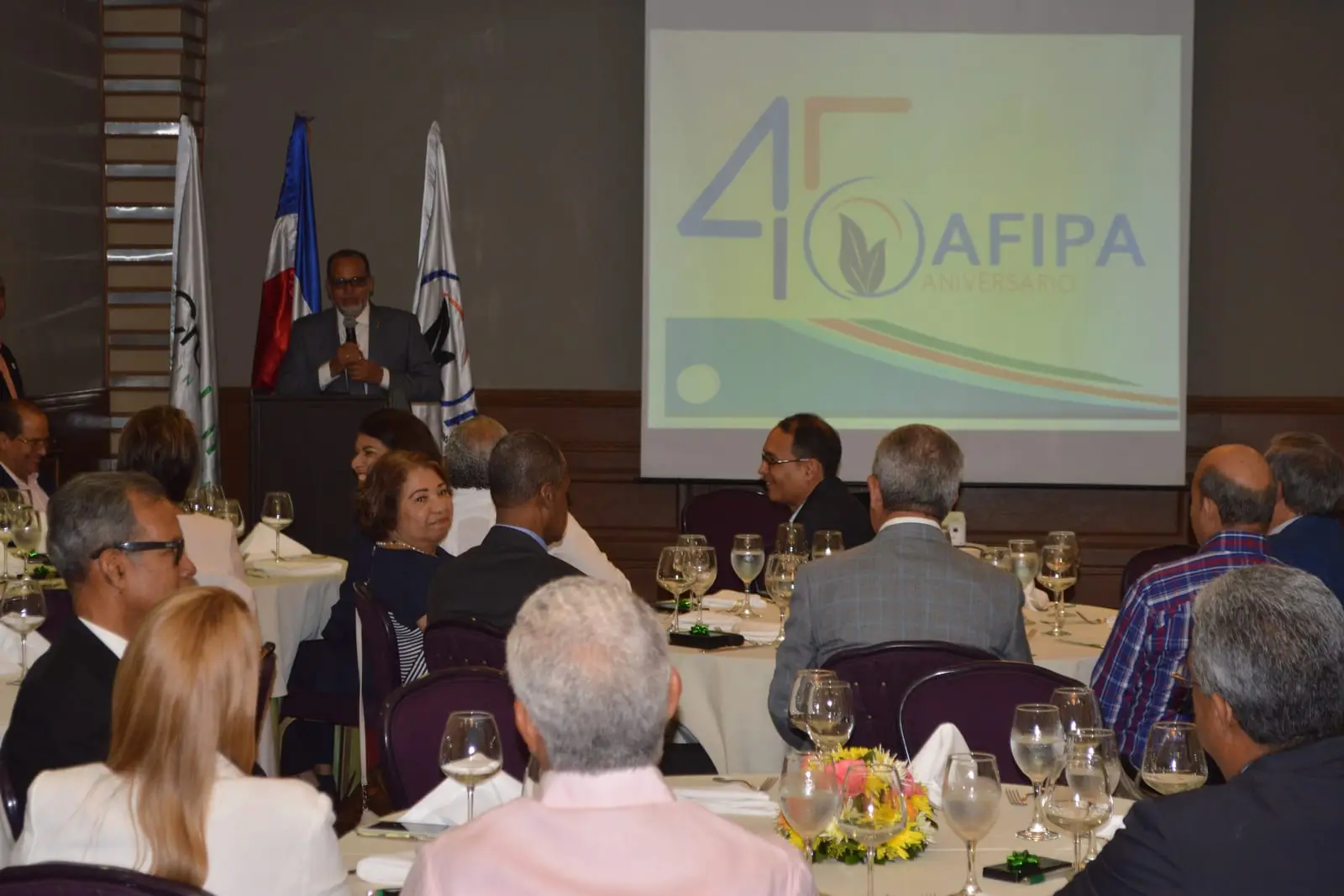 Invitados sentados a las mesas del almuerzo escuchan el discurso, con la gráfica del 45 aniversario de AFIPA proyectada al fondo del salón