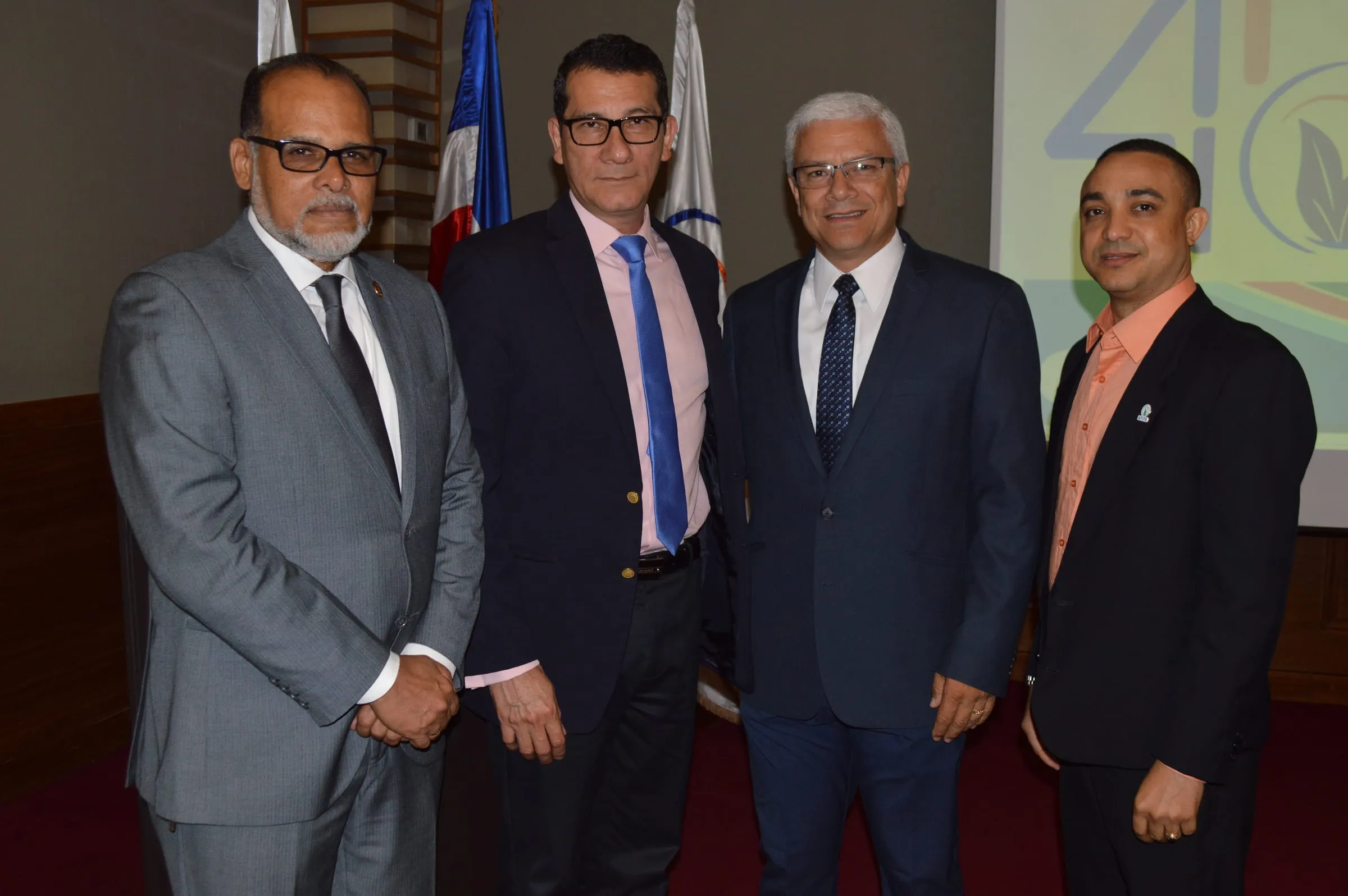 Cuatro directivos e invitados posan juntos en el salón del almuerzo, con las banderas y la gráfica del aniversario detrás