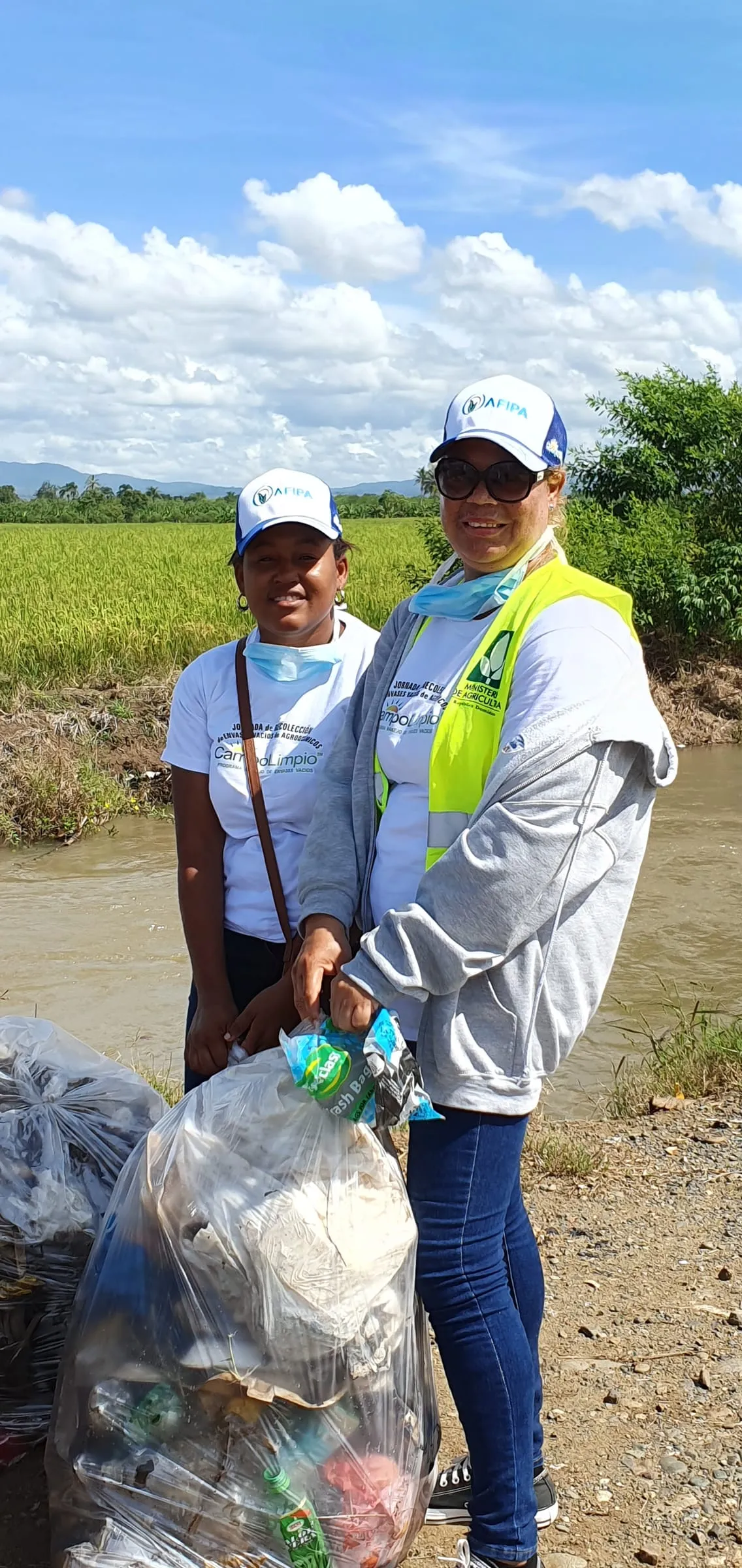 Dos voluntarias con gorra de AFIPA y camiseta de Campo Limpio sostienen una funda llena de envases vacíos frente a un arrozal