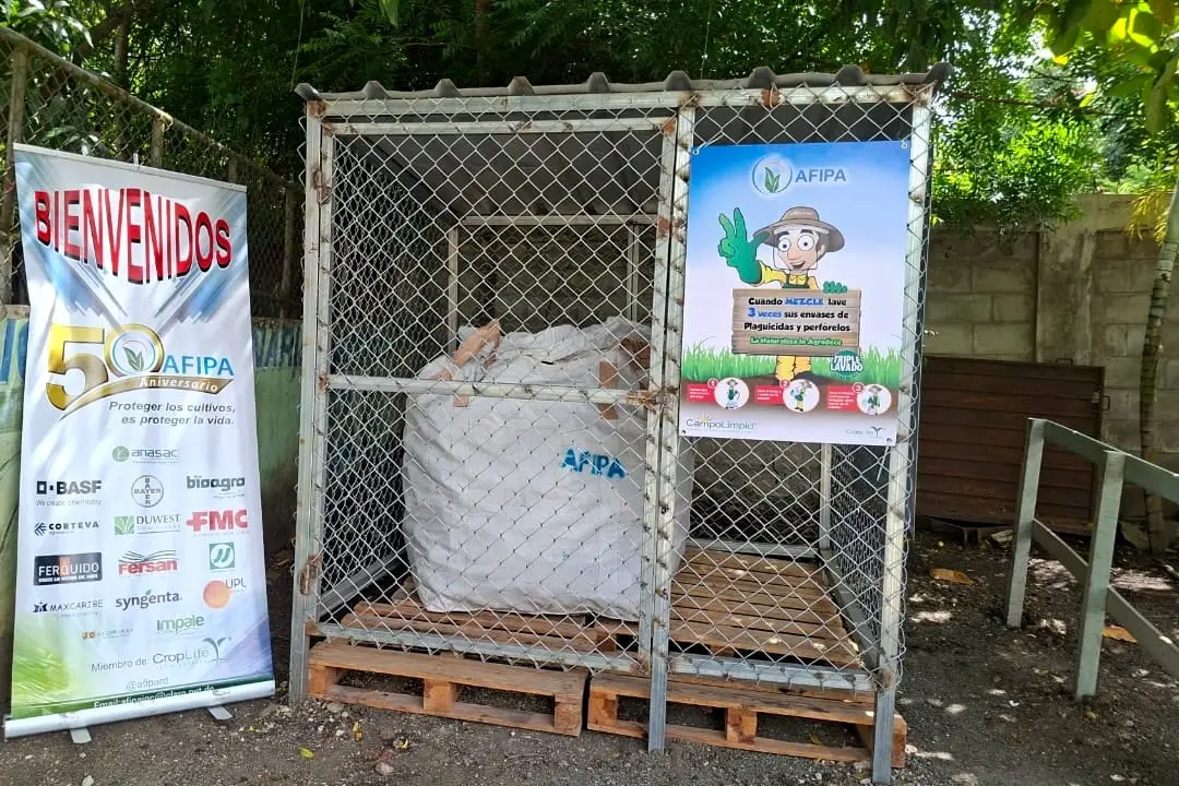 Jaula de malla con un saco de envases vacíos en el centro de acopio de Moca, junto a un letrero de Campo Limpio y un cartel por los 50 años de AFIPA